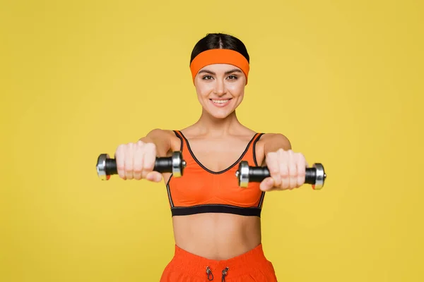 Glückliche Sportlerin schaut in die Kamera, während sie mit auf Gelb isolierten Kurzhanteln trainiert — Stockfoto