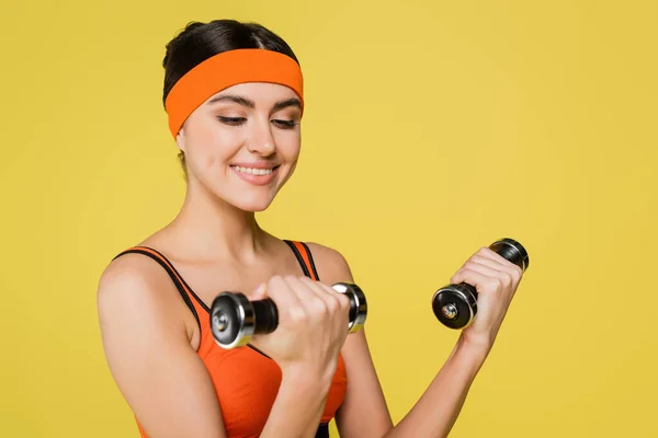 Pleased sportswoman in orange top training with dumbbells isolated on yellow — Stock Photo