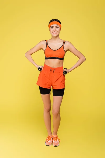 Full length view of sportive woman holding dumbbells while standing with hands on hips isolated on yellow — Stock Photo