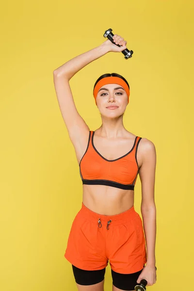 Young sportive woman smiling at camera while exercising with dumbbells isolated on yellow — Stock Photo