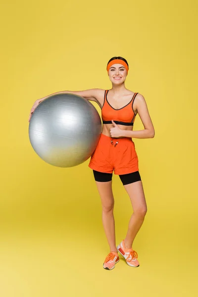 Full length view of happy sportswoman holding fitness ball and showing thumb up isolated on yellow — Stock Photo