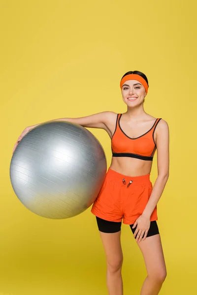Happy sportive woman with silver-grey fitness ball smiling at camera isolated on yellow — Stock Photo