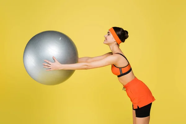 Visão lateral da esportista sorridente exercitando com bola fitness isolada em amarelo — Fotografia de Stock