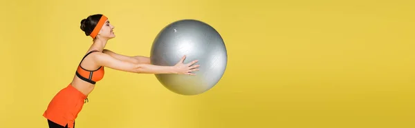 Side view of sportive woman exercising with fitness ball isolated on yellow, banner — Stock Photo