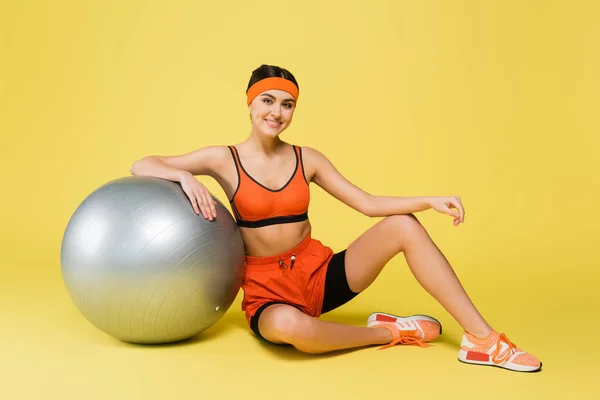 Ziemlich sportliche Frau blickt in die Kamera, während sie in der Nähe von Fitnessball auf gelbem Hintergrund sitzt — Stockfoto