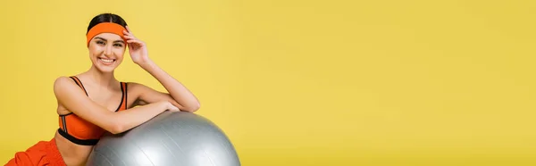 Pretty sportswoman smiling at camera near fitness ball isolated on yellow, banner - foto de stock