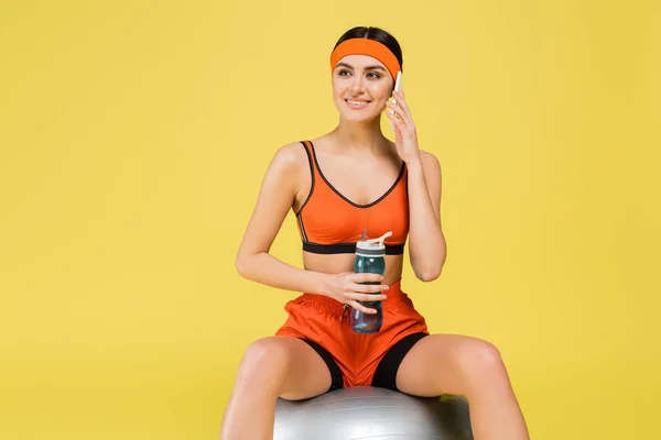 Веселая спортсменка сидит на фитнес-мяч со спортивной бутылкой и разговаривает по сотовому, изолированному на желтый — стоковое фото