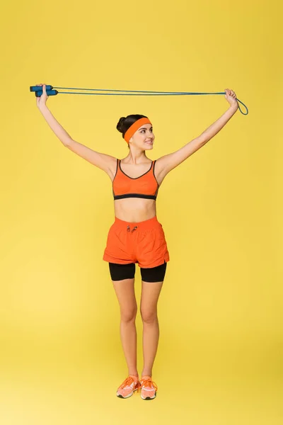 Жінка в помаранчевому спортивному одязі тримає пропускаючу мотузку в піднятих руках ізольовано на жовтому — Stock Photo