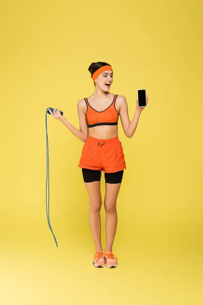 Visão de comprimento total de esportista com corda de salto segurando smartphone com tela em branco isolada no amarelo — Fotografia de Stock