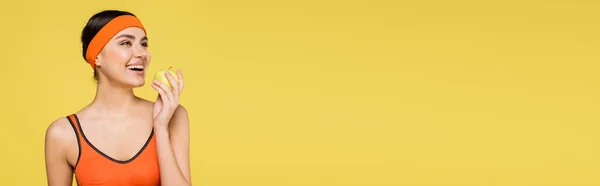 Sportiva sorridente con mela succosa distogliendo lo sguardo isolato sul giallo, striscione — Foto stock