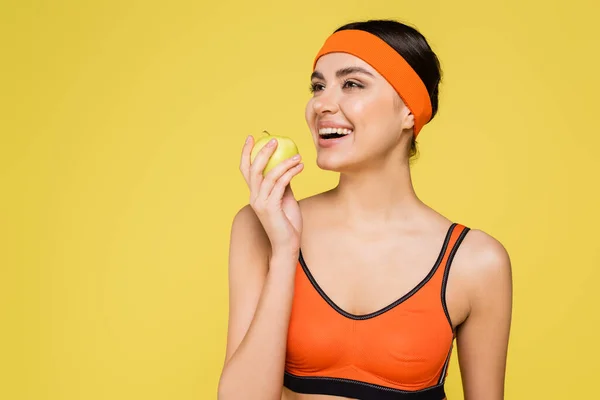 Cheerful sportswoman looking away while holding fresh apple isolated on yellow - foto de stock