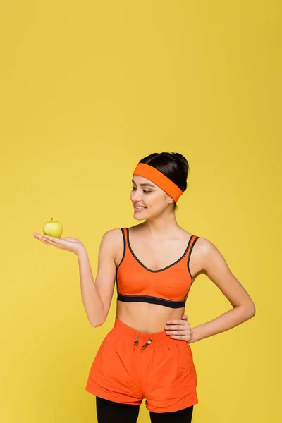 Sportive woman with ripe apple standing with hand on hip isolated on yellow — Stock Photo