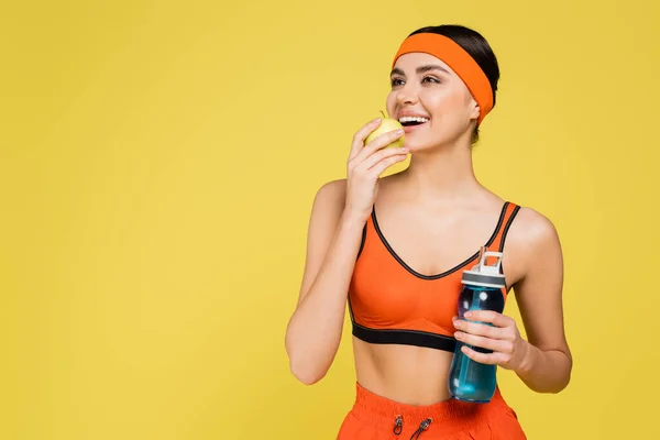 Щаслива спортсменка зі свіжим яблуком і спортивною пляшкою посміхається ізольовано на жовтому — стокове фото