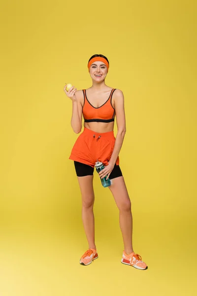 Ganzkörperansicht der jungen Sportlerin mit Apfel und Sportflasche isoliert auf gelb — Stockfoto