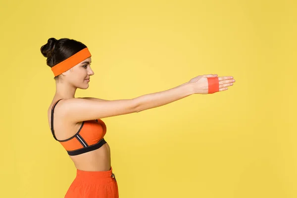Side view of young woman in orange sportswear exercising with resistance band isolated on yellow - foto de stock