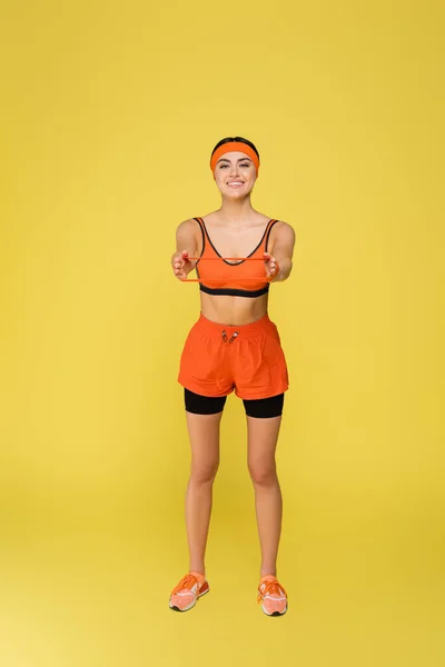 Ganzkörperansicht einer lächelnden Sportlerin, die mit Gummizügen trainiert, die auf gelb isoliert sind — Stockfoto