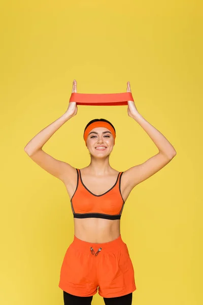 Cheerful woman in orange sportswear training with resistance band isolated on yellow — Stock Photo