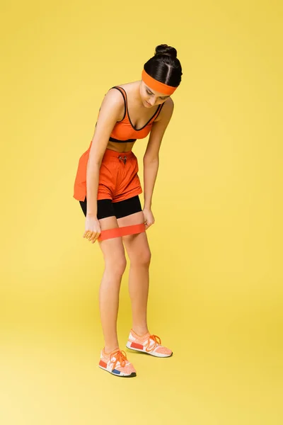 Mulher esportiva colocando banda de resistência nas pernas sobre fundo amarelo — Fotografia de Stock