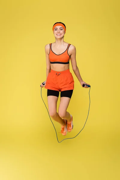 Pleased sportswoman looking at camera while jumping with skipping rope isolated on yellow — Stock Photo