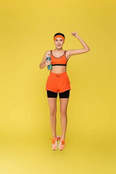 Cheerful sportswoman with sports bottle jumping and showing win gesture isolated on yellow — Stock Photo