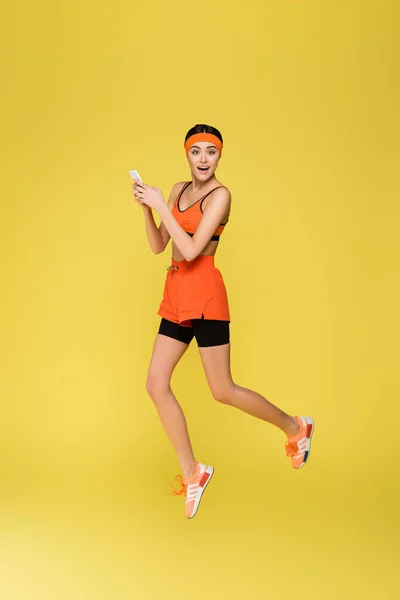 Junge Sportlerin springt beim Smartphone isoliert auf Gelb — Stockfoto