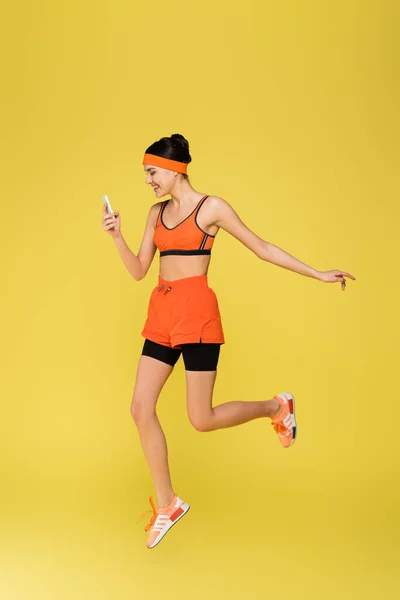 Cheerful sportswoman with smartphone jumping isolated on yellow — Stock Photo