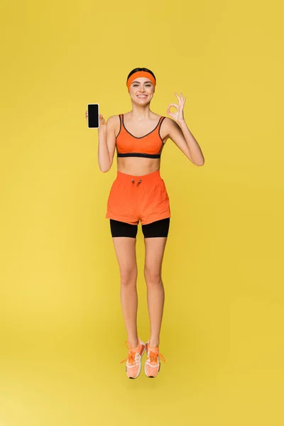 Joyful sportswoman showing okay gesture and smartphone with blank screen isolated on yellow — Stock Photo