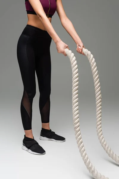 Partial view of sportswoman in black leggings and sneakers training with battle ropes on grey background — Stock Photo