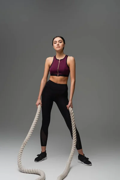 Full length view of woman in black sportswear standing with battle ropes on grey background - foto de stock