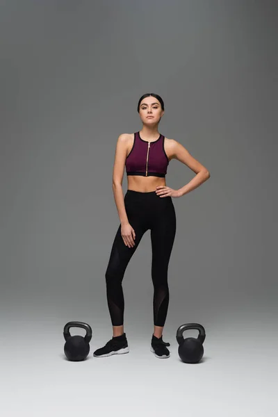 Confident sportswoman standing with hand on hip near kettlebells on grey background — Stock Photo