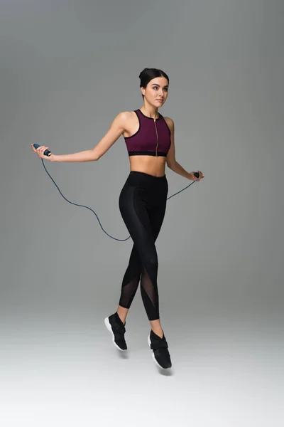 Full length view of sportive woman training with jump rope on grey background - foto de stock