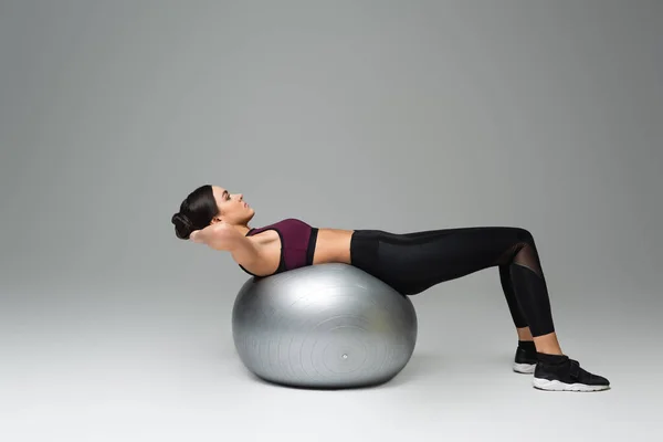 Visão lateral da mulher no treinamento sportswear preto na bola de fitness no fundo cinza — Fotografia de Stock
