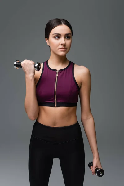 Sportive woman in black top looking away while training with dumbbells isolated on grey — Stock Photo