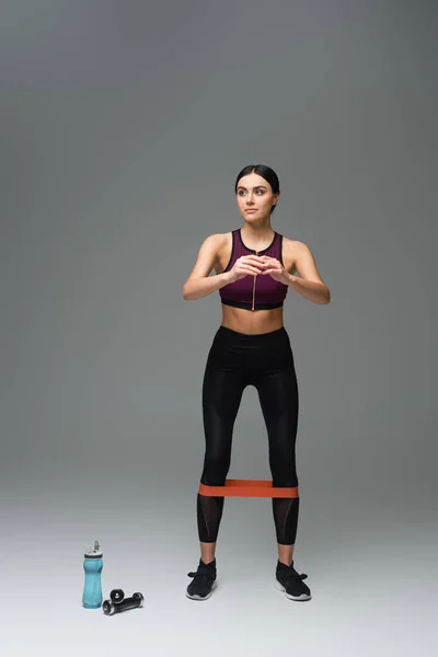 Full length view of sportive woman looking away while training with elastics on grey background - foto de stock