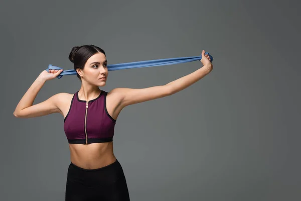 Young sportswoman training with resistance band isolated on grey — Stock Photo