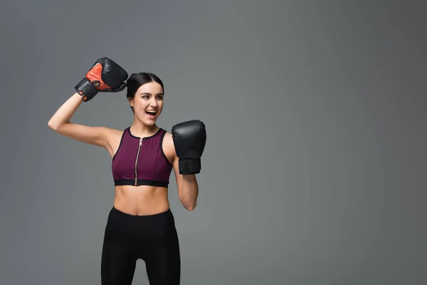 Excited sportswoman in boxing gloves showing win gesture isolated on grey — Stock Photo