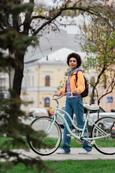 Junge Afroamerikanerin mit Fahrrad und Rucksack blickt auf der Straße in die Kamera — Stockfoto