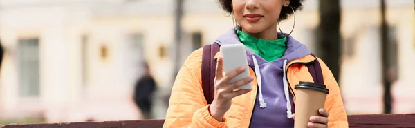 Vista recortada de la joven afroamericana con mochila usando teléfono inteligente y sosteniendo el café para salir al aire libre, pancarta - foto de stock