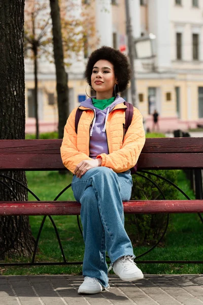 African american woman in jacket sitting on bench on urban street — Stock Photo