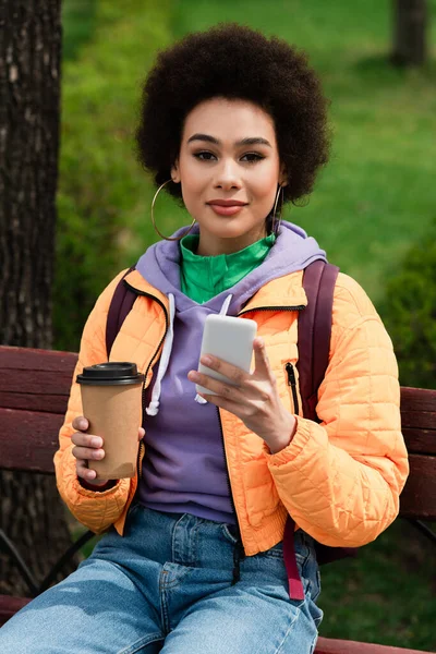 Африканська американка з мобільним телефоном і паперовою чашечкою дивиться на камеру на лавці в парку. — стокове фото