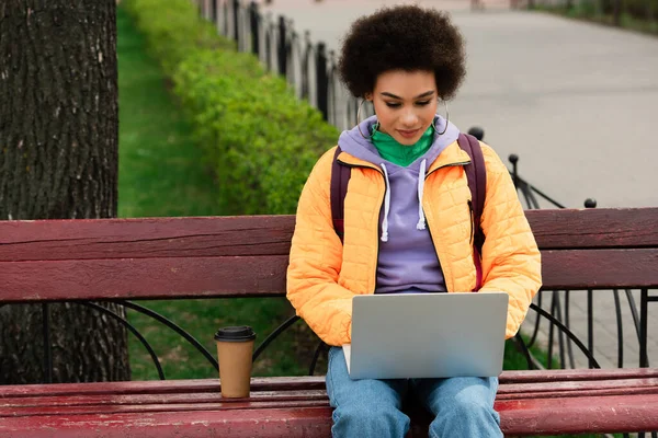 Femme afro-américaine avec sac à dos en utilisant un ordinateur portable près du café pour aller sur le banc — Photo de stock