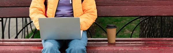 Vue recadrée du télétravailleur utilisant un ordinateur portable près du café sur le banc à l'extérieur, bannière — Photo de stock