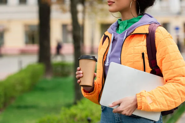 Vista recortada de la mujer afroamericana sosteniendo taza de papel y portátil en la calle urbana - foto de stock