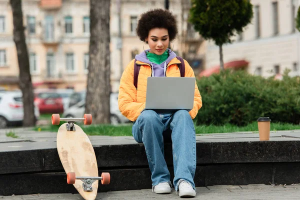African american freelancer using laptop near coffee to go and longboard outdoors — Stock Photo