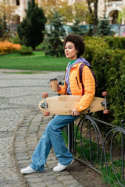 African american woman with paper cup and longboard sitting on fence on urban street — Stock Photo