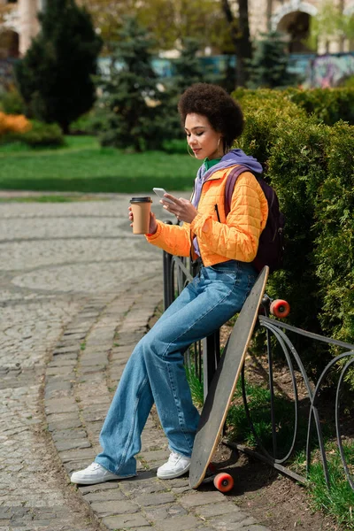 Afroamerikanerin mit Imbissbecher mit Handy in der Nähe von Longboard auf Stadtstraße — Stockfoto