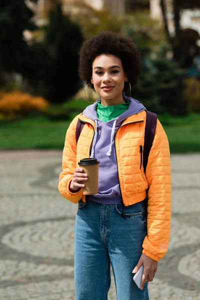 Позитивная африканская американка с мобильным телефоном и бумажной чашкой, смотрящая на камеру на улице — стоковое фото