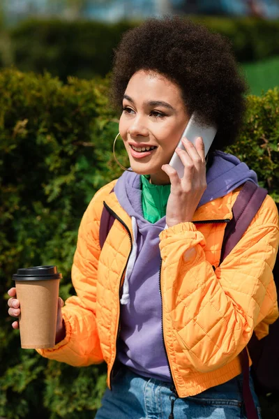 Heureuse femme afro-américaine veste tenant tasse en papier et parlant sur téléphone mobile à l'extérieur — Photo de stock