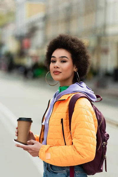 Jeune femme afro-américaine avec sac à dos, tasse en papier et smartphone regardant la caméra à l'extérieur — Photo de stock