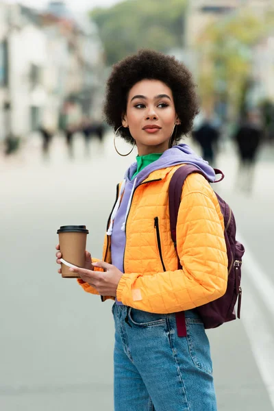 Africano mulher americana com mochila segurando celular e café para ir na rua urbana — Fotografia de Stock